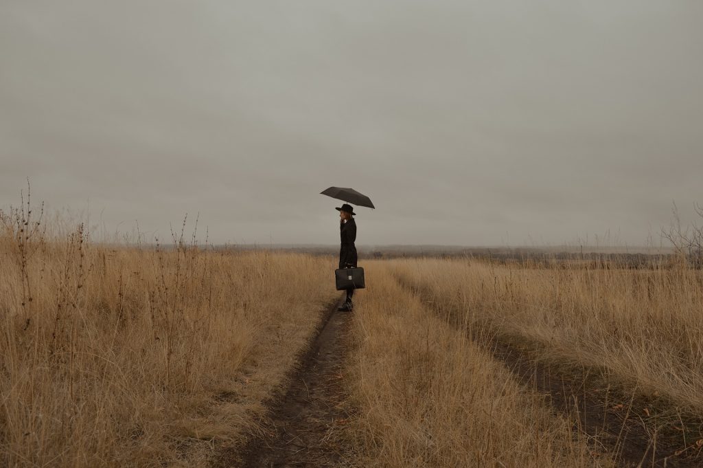 A lady with long skirts and an umbrella, on a long, straight road through prairie grasses.