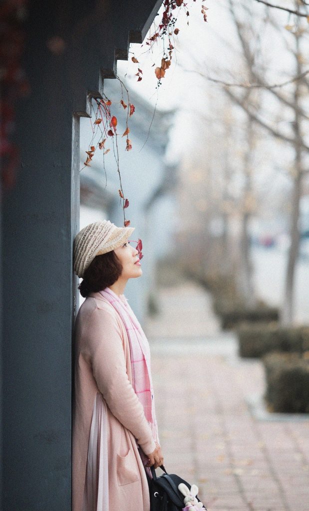 A lady in a pink hat and coat, standing on a tree-lined sidewalk, with her back to the pillar of a building, and her head tilted up to the sky.