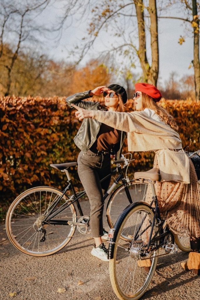 The picture is of two girls with stopped bicycles, one is pointing and the other shading her eyes looking in that direction. One is wearing pants with a denim jacket, t shirt underneath, and black hat. The other is half hidden by the bicycle but appears to be wearing a sweater, skirt, and a red beret. There’s a hedge behind them that’s beside the road, and it’s covered in brownish orange leaves.