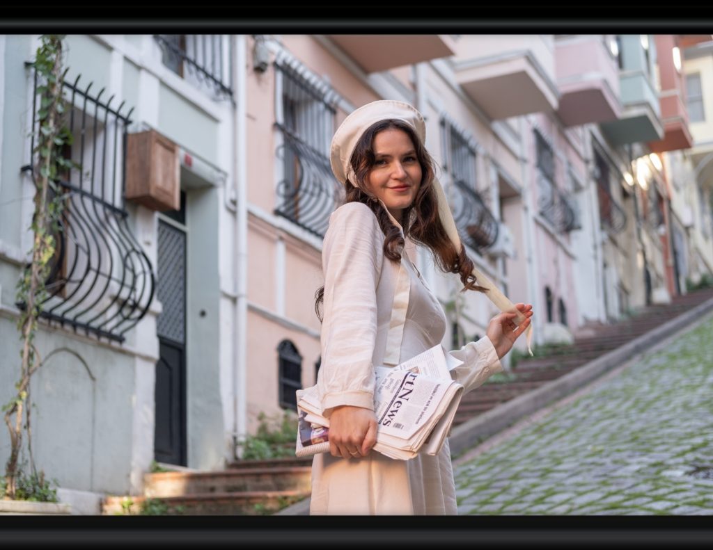 A lady stands on a sloping, European-style street. She is wearing a plain cream-colored dress and a flat cream hat with a tie hanging from each side. She is holding a newspaper in one hand and her hat tie in the other. The street is cobblestoned, and there is grass growing between the cracks. The pastel buildings are all connected and have iron grates over the windows. There is a door every so far. Steps run beside the cobbled road directly in front of the buildings, and there are plants growing on them.