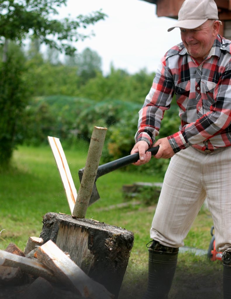 An older man is splitting wood with an ax. The man is wearing a plaid shirt, ballcap, and his pants are tucked into his boots.  He has just brought the ax down, and he is grimacing in a focused way as he looks at the divided piece of wood falling toward the other pieces in the wood pile.