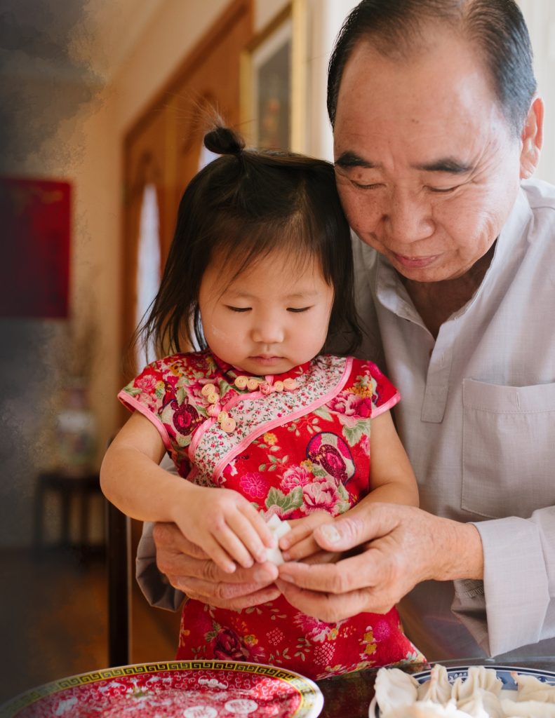 An older Oriental gentleman is helping a toddler shape dumplings. His face has a kind smile, and the little girl is completely focused on what she is doing. The toddler is wearing a floral dress with rosettes.
