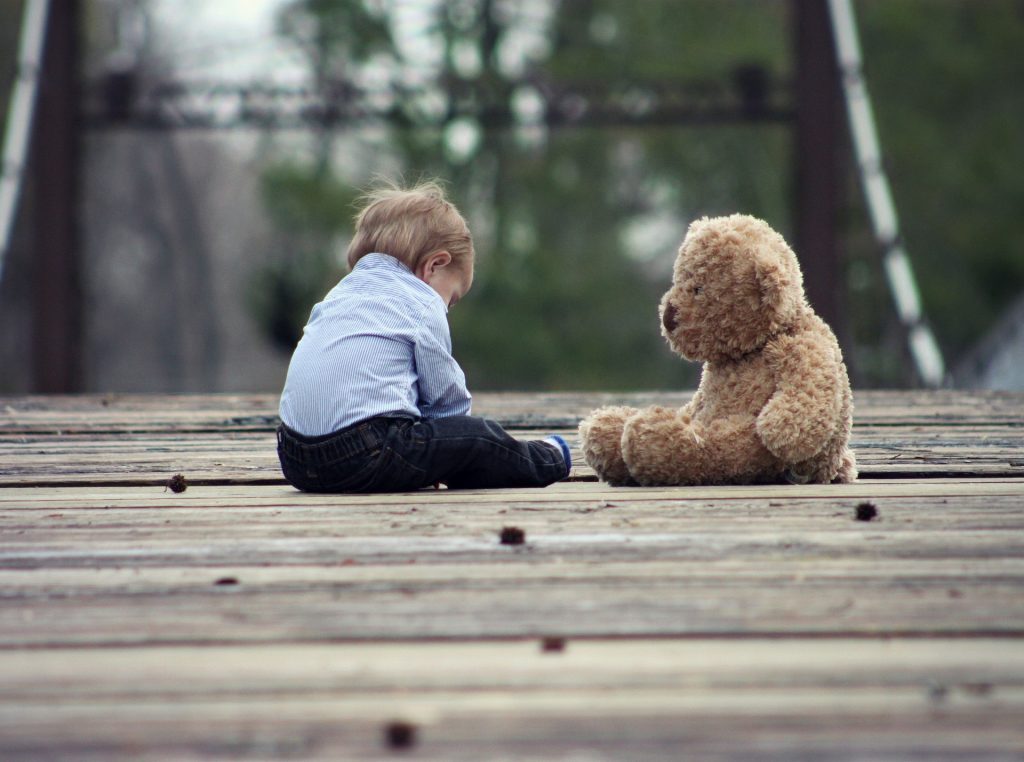 A small toddler boy sits with his back to the camera.  He is wearing a light blue collared dress shirt and very dark blue jeans.  Beside him sits a curly brown teddy bear as big as he is, looking at him like it’s having a conversation with him.  They are seated on a weathered plank walkway  that appears to be a bridge, with trees on the other side.
