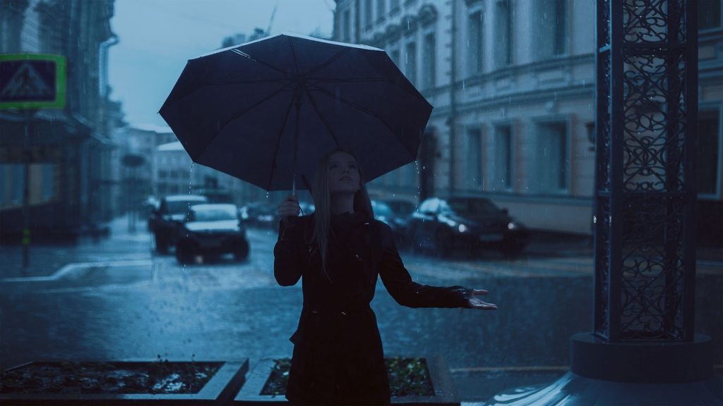 A young woman standing on a sidewalk of a metropolitan street, with large, old buildings towering behind, holding her umbrella and looking upwards, hand outstretched to feel if the rain is still falling.