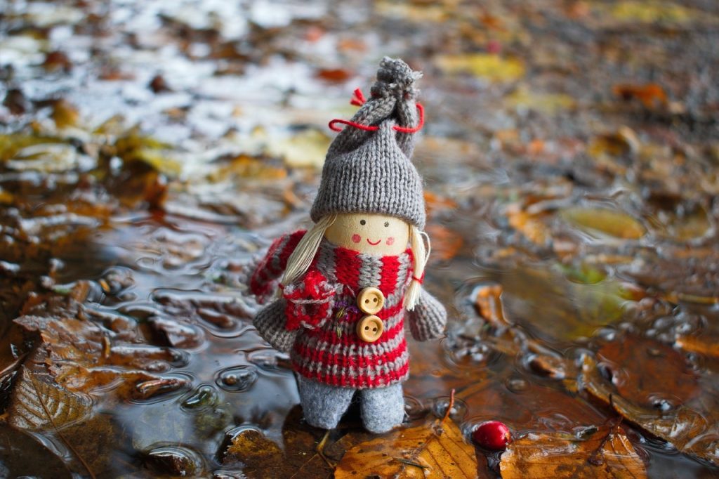 A quaint handmade doll in a knitted red-and-grey outfit with a huge, pompomed hat, standing in a puddle paved with autumn leaves and berries.
