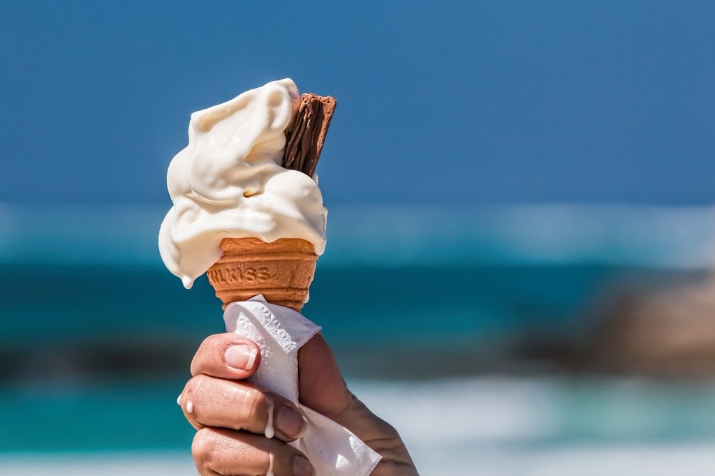 Picture shows a hand holding a melty ice cream cone with a chocolate cookie stuck into the top on one side. The bottom of the cone is wrapped in a napkin, and drips of melted ice cream run down the fingers. The background is heavily blurred, but seems from the colouring to be an ocean, with maybe some rocks or a pier closer to the beach.