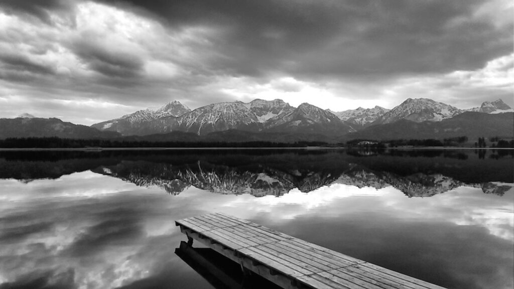 Black and white picture of a narrow dock leading out into a lake with majestic looking mountains in the distance with a cloudy sky.