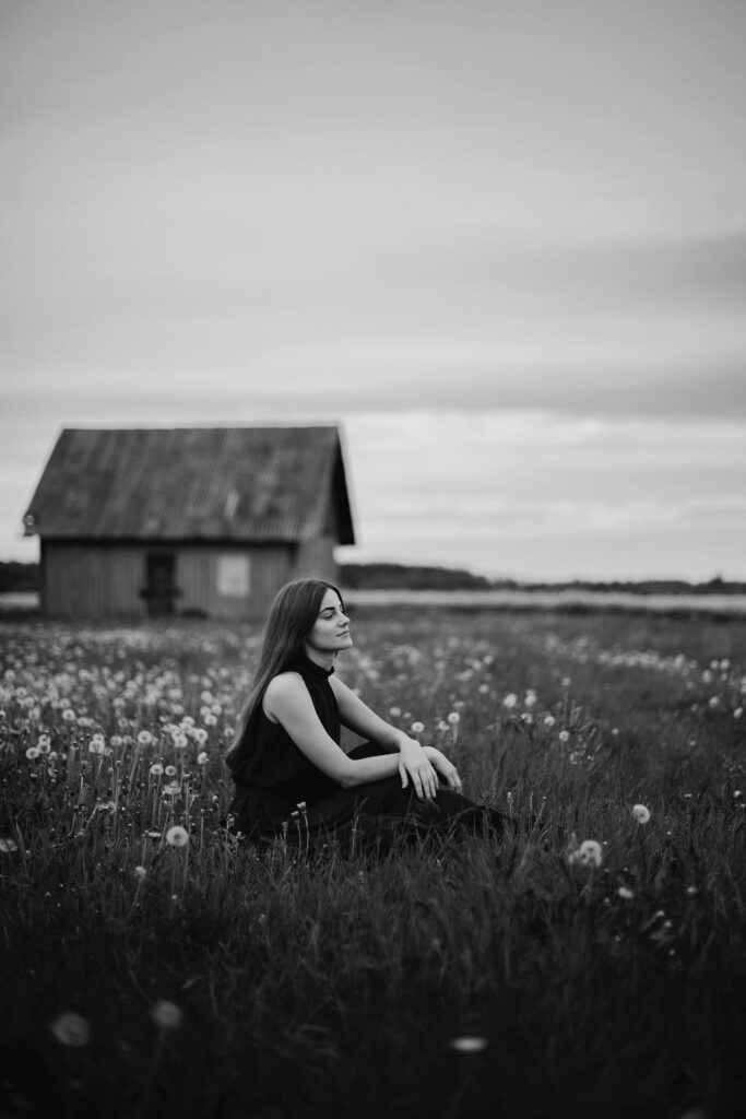 Black and white picture of a girl sitting in a field of flowers, dandelions, I believe, with a small cabin behind her. The sky is also cloudy.