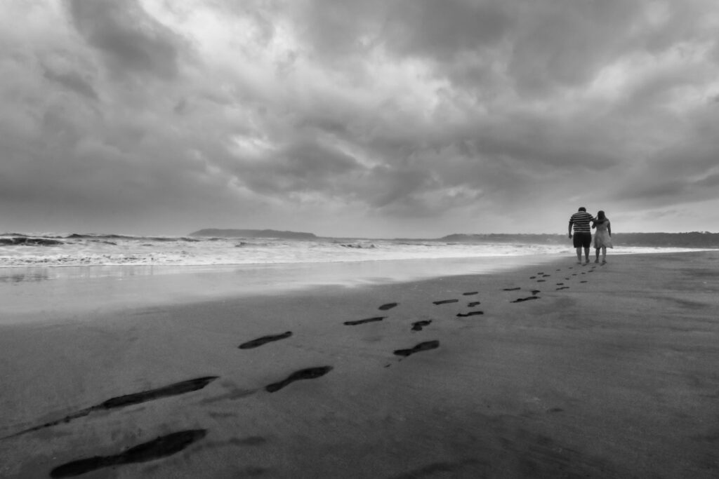 Black and white picture of couple of people walking along the shore at the beach. The sky looks a bit stormy and the ocean is to their left in the picture.