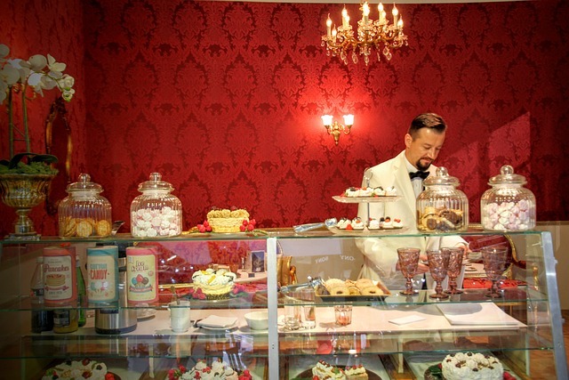 A picture of an old-fashioned candy counter with a man in a white coat standing behind it. The room is lit by a chandelier and a wall sconce, and the wallpaper is covered in a bright red pattern.